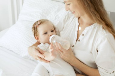 Küçük kızını besleyen mutlu genç anne, yatakta oturup şişeyi tutuyor, boş yer var.
