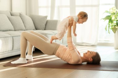 Spor ve annelik kavramı. Evde egzersiz yapan, yoga minderinde uzanan küçük kız bebeğiyle mutlu bir anne.