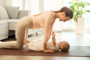 Anne, küçük kızıyla evde egzersiz yapıyor, kız çocuğu yoga minderinde yatıyor, anne ise egzersizler yapıyor, yan görüş yapıyor.