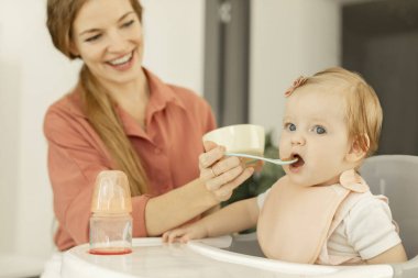 Mutlu anne küçük kızını meyve ya da sebze püresiyle besliyor, bebek mutfakta yüksek sandalyede oturuyor.
