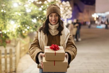 Noel hediyelerini paketleyen mutlu Avrupalı kadın dışarıda kameraya gülümsüyor Noel ışıklarının ve Noel ağacının üzerinde duruyor.