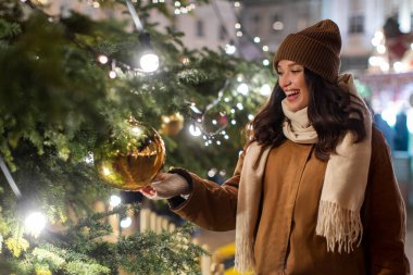 Aşırı mutlu Avrupalı bir kadın süslenmiş Noel ağacında altın topa dokunuyor, Noel tatillerini dışarıda, Noel panayırında geçiriyor.