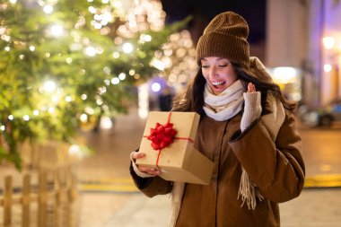 Güzel, sürpriz Avrupalı bir kadın hediye kutusunun açılışını kutluyor, dışarıda süslenmiş Noel ağacının önünde çelenk ışıklarıyla bekliyor.