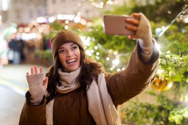 Mutlu bir bayan Noel ağacıyla selfie çekerken gülüyor arka planda ise el sallıyor, telefona gülümsüyor ve dışarıda ailesiyle görüntülü konuşma yapıyor.