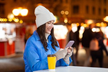 Kahve fincanı olan mutlu genç bir kadın Noel pazarında cep telefonu kullanıyor, bayram geçmişi, boş yer.