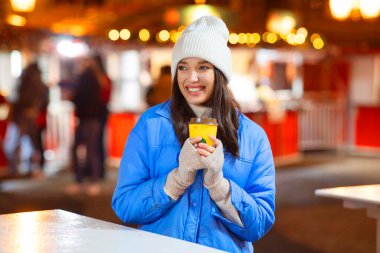 Açık havada sıcak sıcak şarap içen mutlu bir kadının portresi ve Noel pazarının şenlikli arka planı.