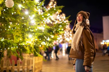 Mutlu genç bir kadın Noel pazarında yürüyor, süslenmiş Noel ağacının önünde çelenk ışıklarıyla duruyor.
