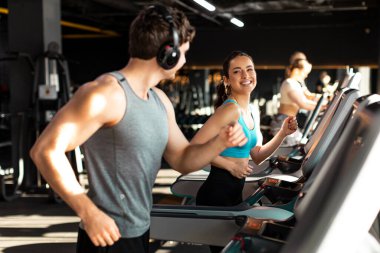 Modern spor salonunda kardiyo antrenmanı yapan mutlu bir adam ve kadın koşu bandında koşan ve birbirleriyle konuşan bir çift.