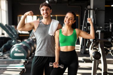 Güçlü bir çift, spor yaptıktan sonra modern spor salonunda poz veren erkek ve kadın, kaslarını gösterip kameraya gülümsüyorlar.