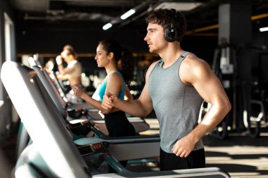 Genç Avrupalı kaslı adam koşu bandında koşuyor ve müzik dinliyor, modern spor salonunda kardiyo çalışması yapıyor.