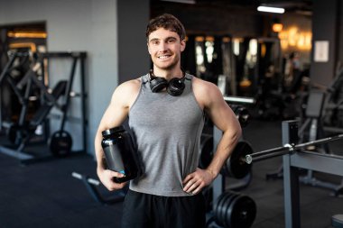 Spor beslenmesi. Siyah kabın içindeki protein sümsük tozlu kaslı adam spor salonunda kameraya gülümsüyor.