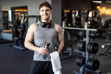 Sportif genç adam antrenmandan sonra spor salonunda duruyor, idmandan sonra su şişesi ve havlu tutuyor, kameraya poz veriyor ve gülümsüyor.