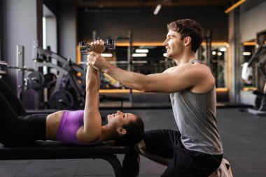 Kişisel antrenör, kadının göğüs ve omuz gerdirme hareketlerinde yardımcı oluyor, erkek antrenör ona yardımcı oluyor ve talimatlar veriyor, yan görüş.