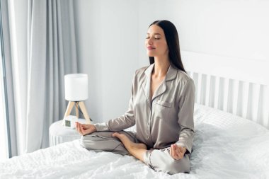 Serene genç kadın beyaz yatakta bağdaş kurarak oturuyor, gözleri kapalı ve elleri meditasyon pozunda.