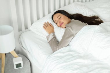 Genç Avrupalı bir bayan beyaz rahat bir yatakta uyuyor, yakınlardaki masada çalar saat, yatak odasının içinde. Sağlıklı uyku, uyanma zamanı, hafta sonu sabahı.