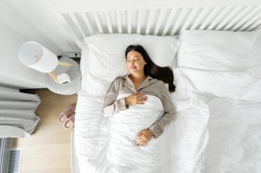 Huzurlu genç bayan sırt üstü uzanıyor, yumuşak yastıkta rahat beyaz bir yatakta uyuyor, battaniyeyle örtülmüş olarak dinleniyor ve üst kattaki manzaranın keyfini çıkarıyor.