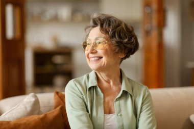 Happy dreamy senior woman in glasses look aside into distance deep in pleasant memories, sitting on sofa at home clipart