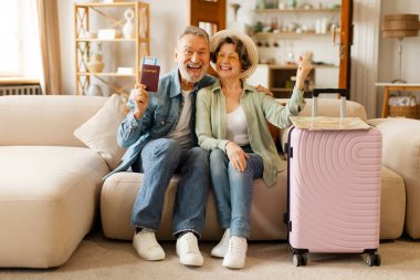 Neşeli Avrupalı yaşlı çift seyahat biletleri ve bavullarıyla poz veriyor, tatil için hazırlanıyor, evdeki kanepede oturuyorlar.