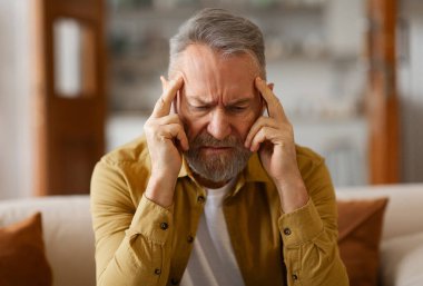 Yaşlı adam şiddetli baş ağrısı çekiyor, şakaklarına dokunuyor, gözleri kapalı kanepede oturuyor, migren ya da gerilim sorunu yaşıyor.
