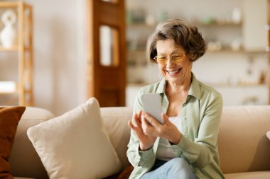 Cep telefonunda cep telefonu uygulamaları kullanan, mesaj yazan, oturma odasındaki koltukta oturan, elinde akıllı telefon tutuyormuş gibi gülümseyen mutlu son sınıf öğrencisi kadın.