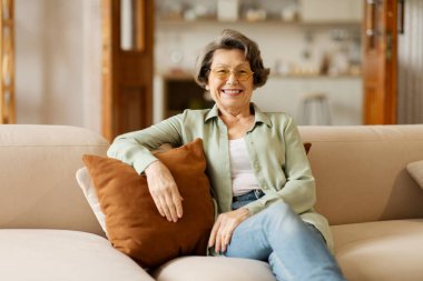 Kanepede oturan, yastığa yaslanan, kameraya bakan ve gülümseyen, rahat bir evde dinlenen son sınıf güzeli kadın.