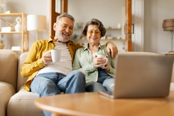 Расслабленная пожилая пара смотрит кино на ноутбуке, отдыхает дома на диване и пьет чай, пожилые мужчины и женщины наслаждаются домашним отдыхом