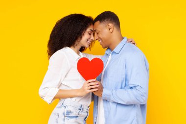 Sarı arka planda sevgililer gününü kutlayan mutlu Latin aşıklar, kucaklaşmalar ve dokunaklı alınlar, kırmızı kalp kartlarını bir arada tutarlar.