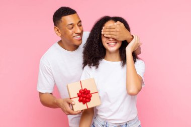 Bayanların gözlerini kapayan, sürpriz yapan, hediye kutusu veren, pembe stüdyo geçmişine karşı birlikte duran bir çift.