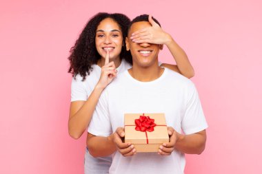 Sürpriz hediye. Latin bayan erkek arkadaşının gözlerini kapatıyor ve sessizlik işareti gösteriyor, pembe arka plan için hediye kutusu veriyor. Sevgililer günü, doğum günü, yıldönümü kutlaması
