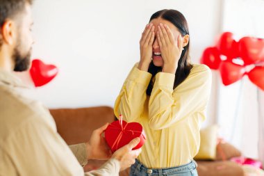 Sevgililer Günün kutlu olsun. Adam sürpriz yapıp, karısına kalp şeklinde hediye paketi verirken kadın elleriyle gözlerini kapatıyor.