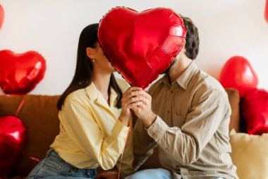 Aşık bir çift kalp şeklinde balon tutuyor, öpüşürken üzerlerini örtüyorlar, evde süslü bir odada oturuyorlar.