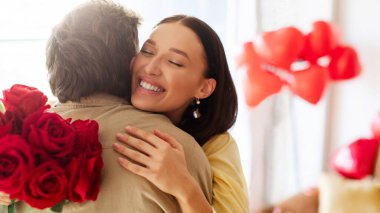 Romantik Sevgililer Günü. Kırmızı gül buketi tutan mutlu kadın ve şefkatle sarılan adam, evde kutlamanın tadını çıkarıyor.