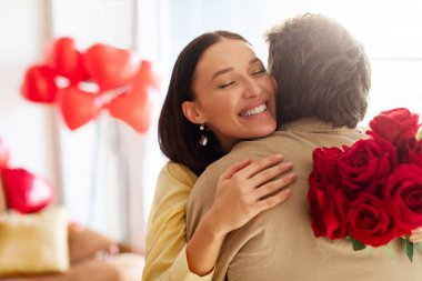 Romantik Sevgililer Günü. Kırmızı gül buketi tutan mutlu kadın ve şefkatle sarılan adam, evde kutlamanın tadını çıkarıyor.