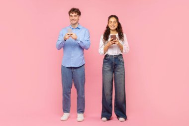 Akıllı telefonlarını kullanan, mesajlaşan, pembe stüdyo arka planında tek başına duran pozitif kadın ve erkeğin vücut uzunluğu.