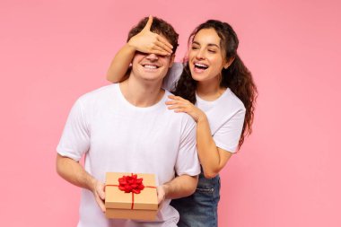 Güzel Avrupalı kadın sevgilisinin gözlerini kapatıyor, ona hediye paketi veriyor ve sevgililer günü, doğum günü ya da yıldönümü kutlamaları yapıyor, pembe stüdyo arka planının başında duruyor.