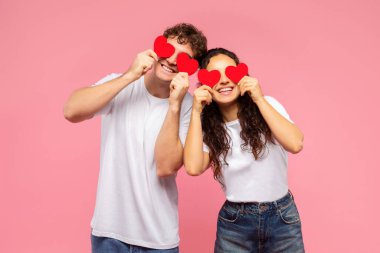 Aşıklar aşkları tarafından kör edildi. Gözlerinin üzerinde kırmızı kalp şeklinde kartlar tutan mutlu bir çift ve gülümseyen, pembe arka planda duran. Sevgililer Günü konsepti