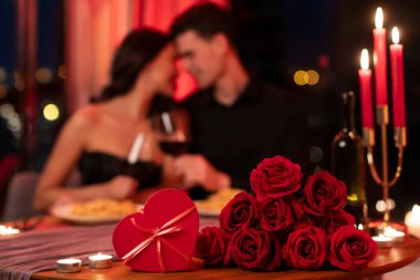 Romantik Sevgililer Günü yemeği. Arka planda birbirine aşık bir çift, akşam vakti atmosferik restoranda buluşuyorlar. Çiçeklere ve hediye kutusuna odaklan
