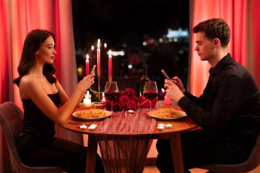 Kötü randevu konsepti. Akıllı telefonlar kullanan sıkıcı bir adam ve kadın, restoranda romantik bir akşam yemeğinde birbirlerini görmezden geliyorlar, masada yan gözle oturuyorlar.