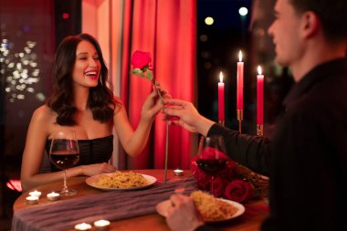 Sevgili koca, yemekte mutlu bir eşe kırmızı gül veriyor. Sevgililer gününü restoranda romantik bir ortamda kutluyor.