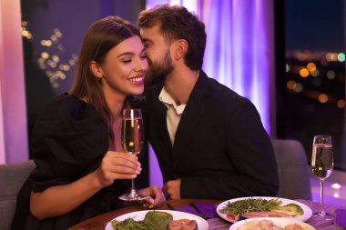 Lokantada yemek yiyen, sevgililer gününü birlikte kutlayan, sevgililerine fısıldayan ve gülümseyen bir kadın.