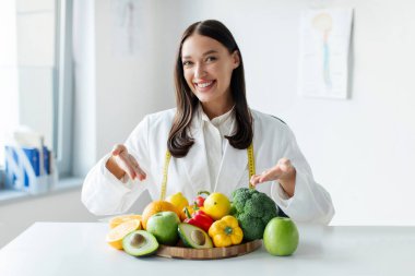 Laboratuvar önlüklü neşeli diyetçi masadaki taze meyve ve sebzeleri işaret ediyor. Kilo kaybı danışmanı sağlıklı bitki temelli diyet öneriyor