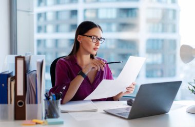 Genç bayan muhasebeci faturaları kontrol ediyor, satış istatistik yönetimini, mali veri belgelerini ya da ofis raporlarını analiz ediyor.