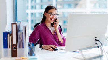 Mutlu genç iş kadını yönetici, cep telefonuyla konuşuyor, iş yerinde iş görüşmesi yapıyor, iş yerindeki modern bilgisayarı kullanıyor.