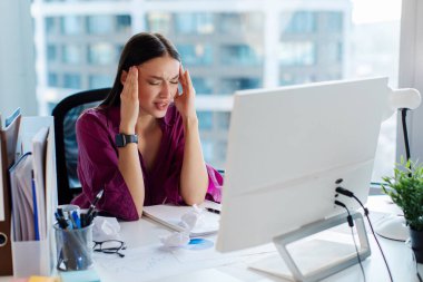Ofiste baş ağrısından muzdarip çok çalışan genç bir iş kadını bilgisayar monitörünün önünde oturuyor, işiyle ilgili sorunlar yaşıyor.