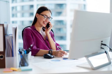 Kadın yönetici cep telefonuyla konuşuyor ve not alıyor, ofisinde masasında oturuyor, bilgisayarla görüşmelere cevap veriyor.