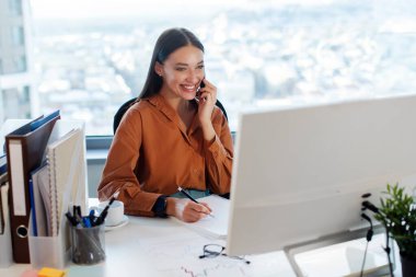 İş kadını cep telefonuyla konuşuyor, bilgisayar ekranına bakıyor ve not alıyor, modern ofiste çalışıyor. Girişimcilik ve iş kariyeri