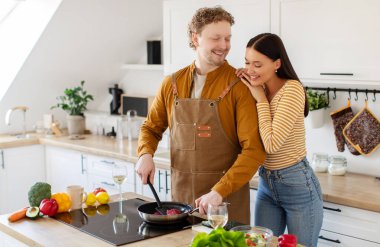 Yakışıklı adam lezzetli yemekler hazırlıyor, mutlu kadın yakınlarda duruyor ve kocasına sarılıyor, mutfak içi, fotokopi odası.