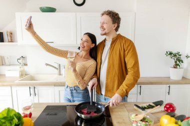 Birlikte yemek hazırlarken cep telefonuyla selfie çeken mutlu çift ve elinde şarapla kadeh tutan kadın evde eğleniyor.