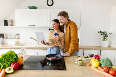 Mutlu çift yemek pişirirken mutfaktaki rahat evde dizüstü bilgisayar ve kredi kartı kullanıyor. Evde online alışveriş.