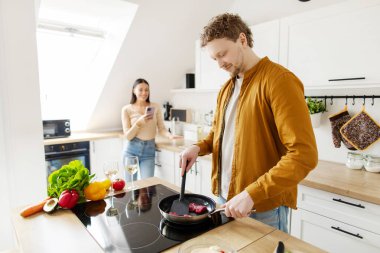 Yakışıklı bir adam akşam yemeği için et pişirirken karısı arka planda cep telefonu kullanıyor.